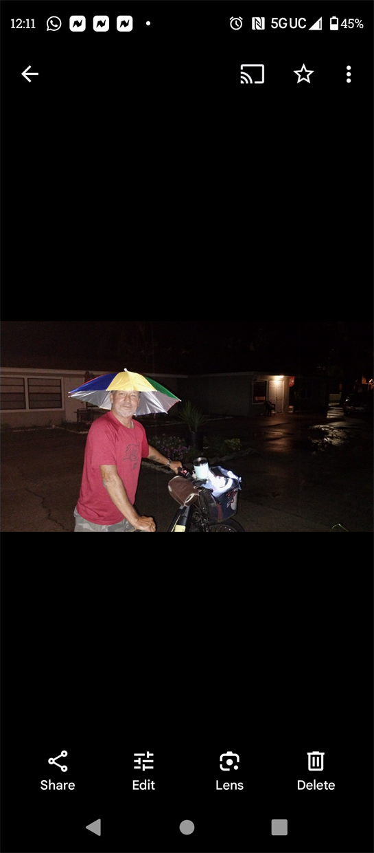 A man is cycling wearing a colorful umbrella hat under the rain at night.