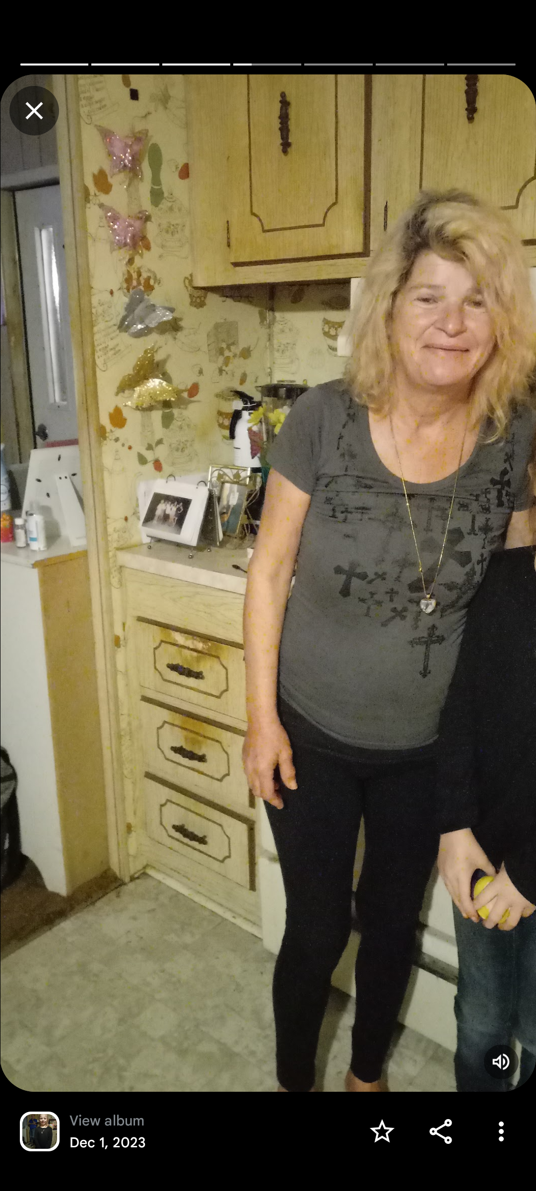 Woman in gray shirt poses beside a colorful wall decorated with family memories.