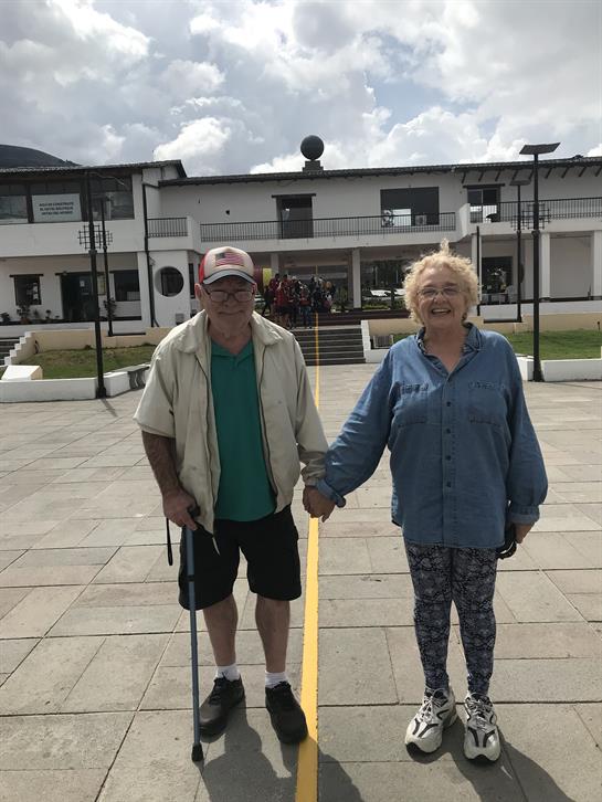 An elderly couple holds hands in front of a community center under a partly cloudy sky.