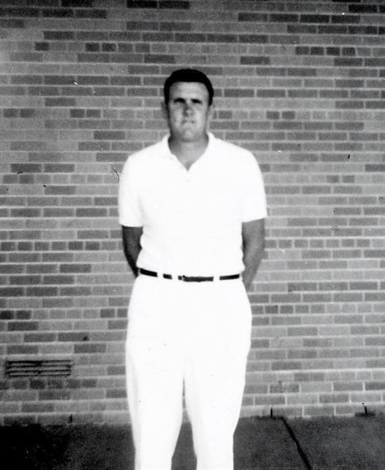 A young man dressed in a white outfit stands with hands behind back against a brick wall.