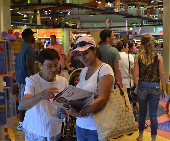 Two women engage in conversation while looking at a magazine in a busy retail environment.