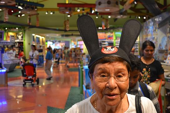 An elderly woman smiles in bunny ears at a lively entertainment center filled with people.