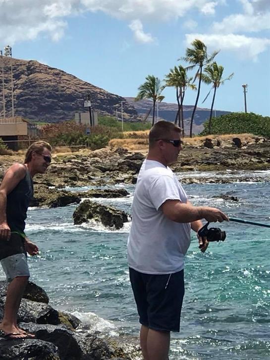 Two men are engaged in fishing on a rocky beach with palm trees in the background.