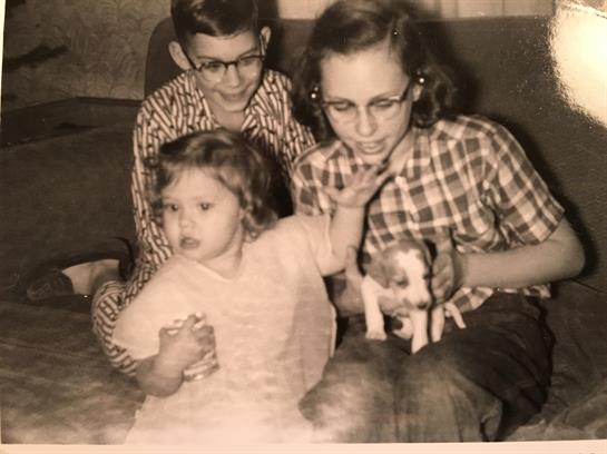Three children sit together in a living room, one holding a small puppy and others playing.