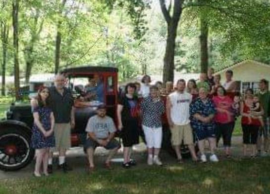 A large group of people enjoys a family reunion by a vintage truck surrounded by greenery.