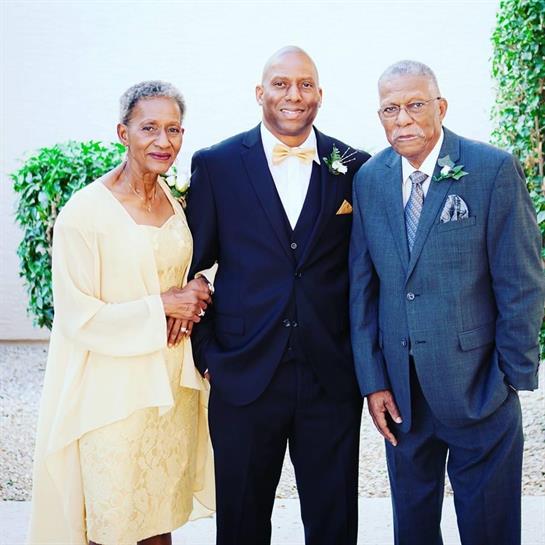 A groom in elegant attire poses with two elderly relatives in a garden filled with greenery.