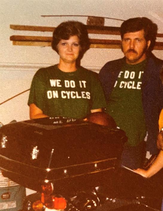 A couple stands proudly beside a motorcycle wearing matching green shirts with fun messages.