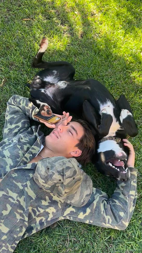 A person lies on soft grass enjoying time with a friendly dog, both appearing relaxed and happy.