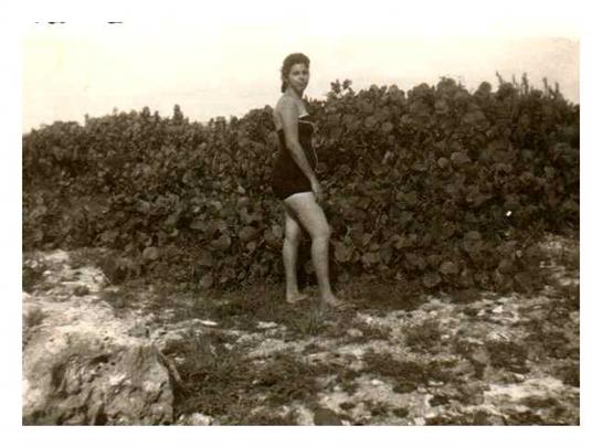A young woman poses in a swimsuit near vibrant greenery at a beach, with clouds overhead.