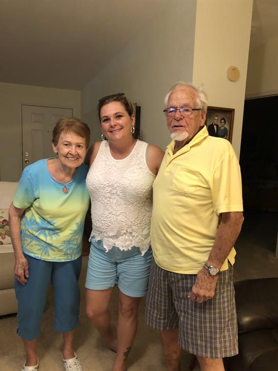 A middle-aged woman stands between an older man and woman, all smiling warmly indoors.