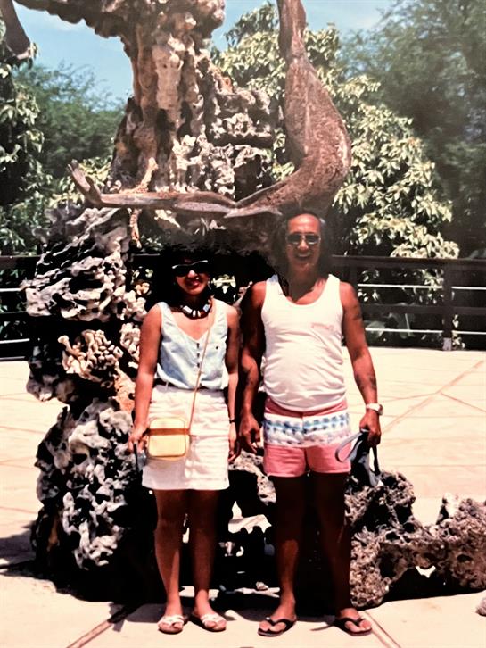 A couple smiles in a lush garden near a striking stone sculpture under clear skies.