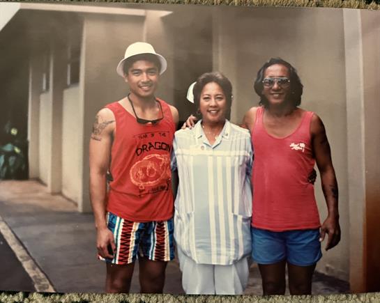 Three individuals dressed in summer outfits stand together, smiling under bright sunlight.