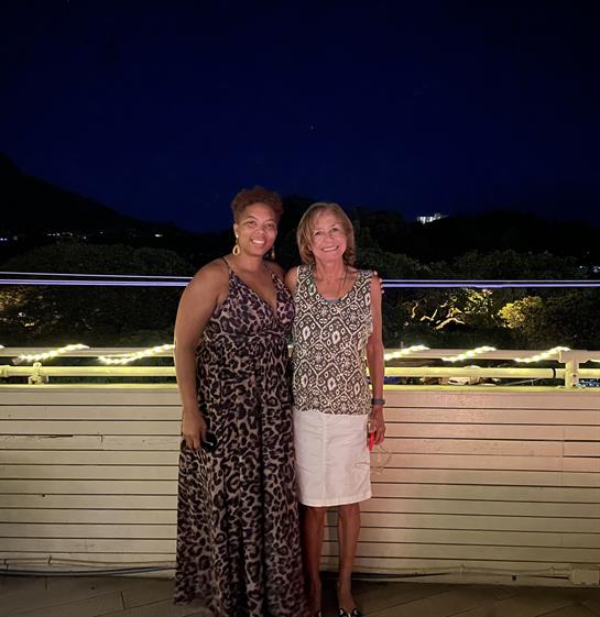 Two women pose happily on a terrace with soft lighting and a darkening sky behind them.