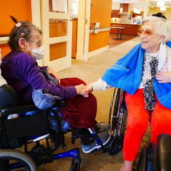 Two elderly women in wheelchairs engage warmly during a visit at a nursing home facility.