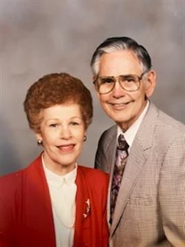 A cheerful couple stands side by side, dressed in formal attire, smiling warmly at the camera.