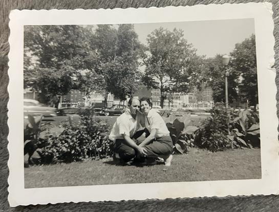 A couple poses happily in a lush garden with trees and flowers on a sunny day.