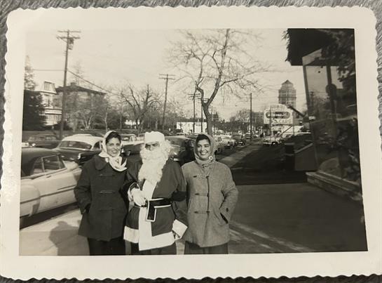 Friends stand outdoors with Santa in winter attire as cars pass by on a lively street.
