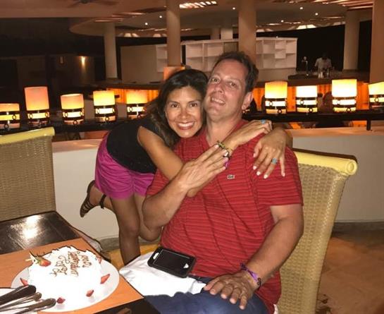 A couple smiles and embraces at a dining table, surrounded by a lively resort atmosphere.