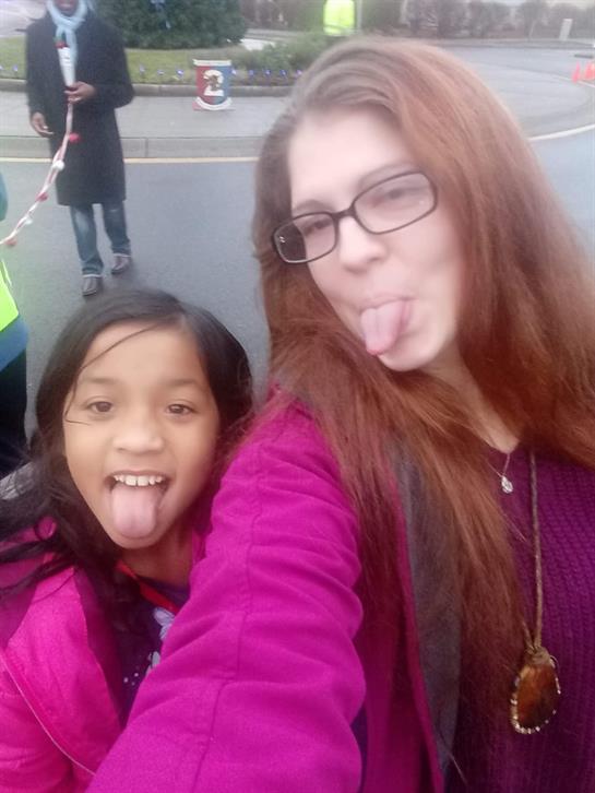Two girls make funny faces for a selfie while enjoying time together outside with others nearby.