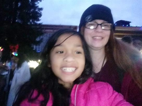 A girl wearing a pink jacket poses happily with a woman at a vibrant outdoor celebration.