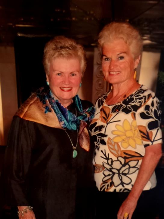 Two women pose happily together in formal attire at a social gathering, smiling warmly.