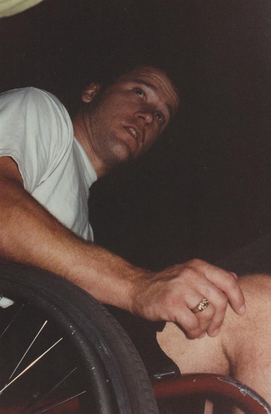 A man sits in a wheelchair, appearing contemplative in a dark setting at night.