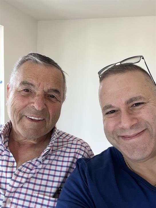 Two men share a smile together while standing in a well-lit room with a neutral background.
