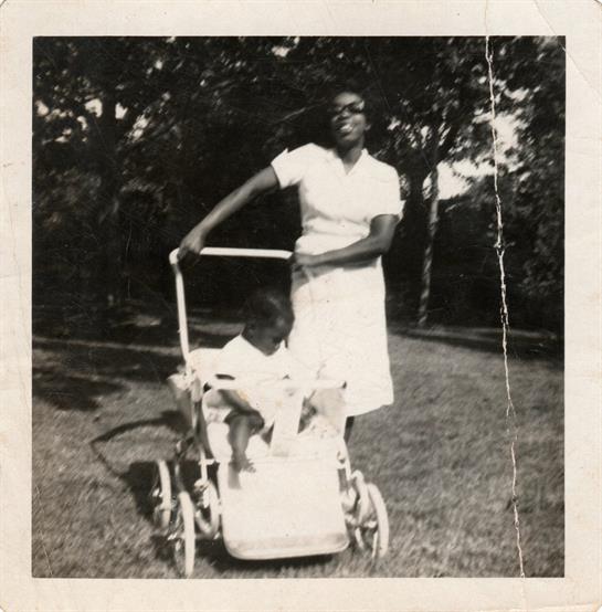 A mother joyfully walks with her child in a stroller in a lush park on a warm day.