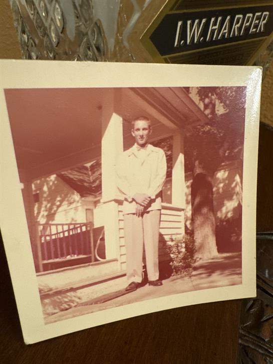 A man poses confidently on a porch in front of a house surrounded by trees.