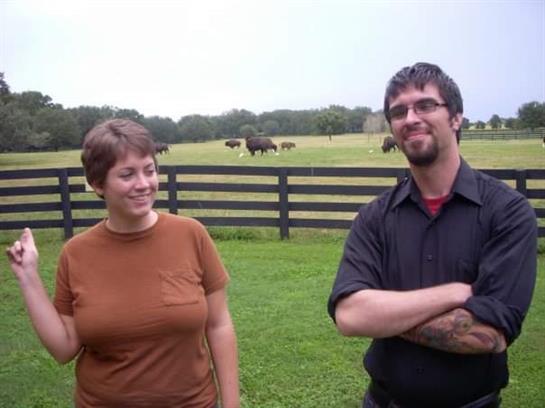 Two people stand on lush green grass, smiling and enjoying a sunny day while bison graze nearby.