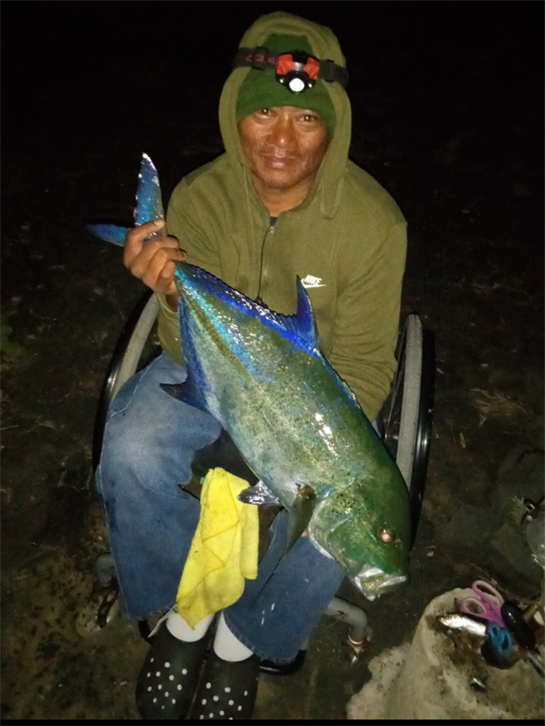 A man in a hoodie shows off a vibrant blue-green fish caught at night by the shore.