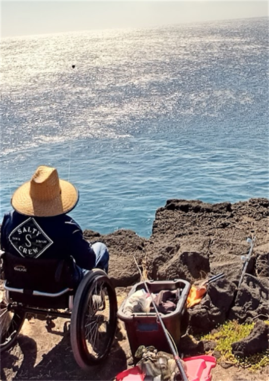 Individual enjoys fishing from rocky area near the ocean while seated in a wheelchair.