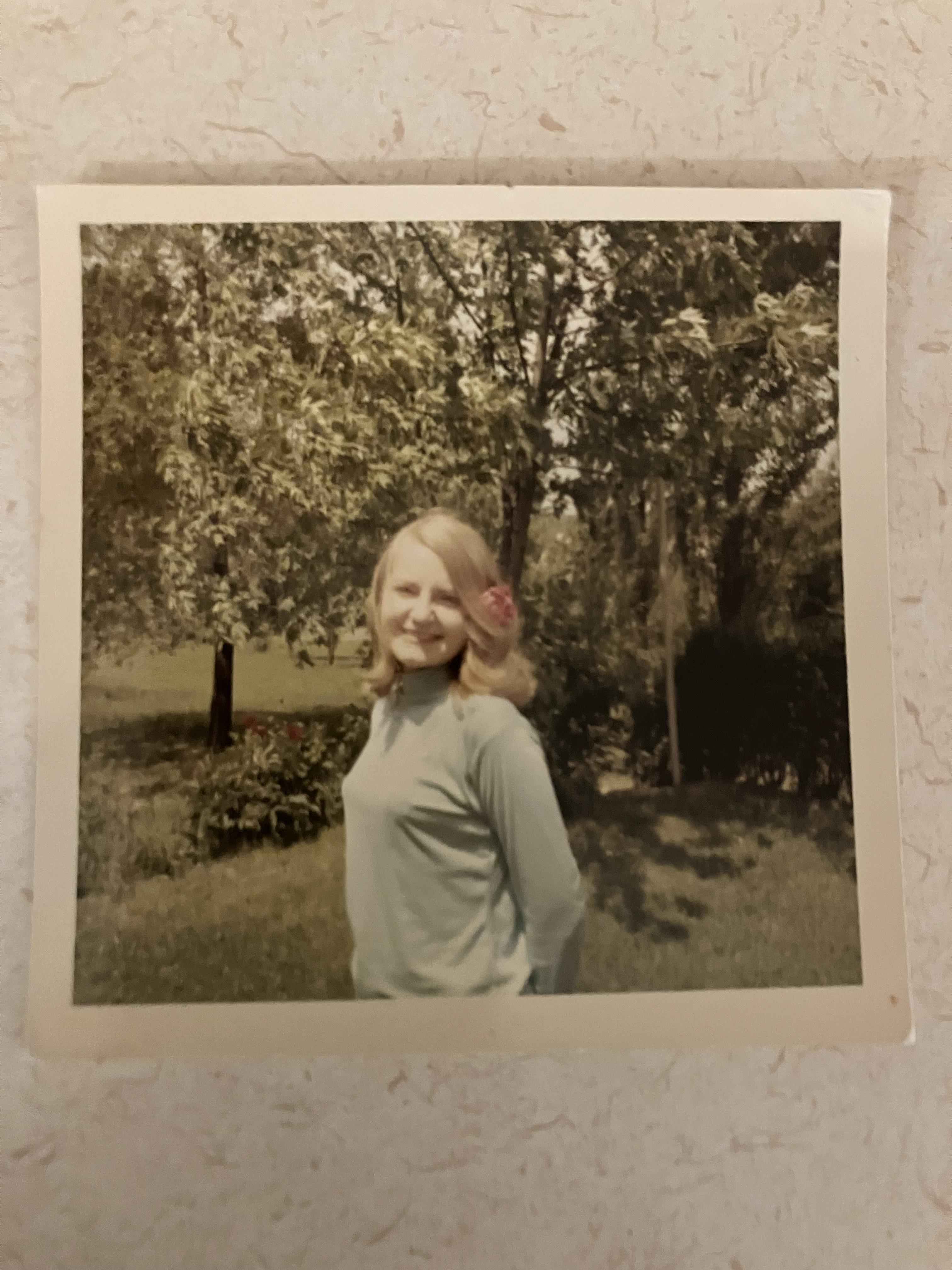 Woman with light hair smiles confidently while standing in a lush, green garden during the day.