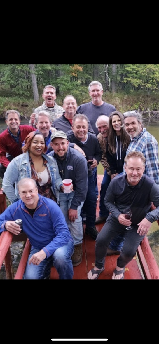 Friends gather on a red dock by a lake surrounded by trees, smiling and holding drinks.