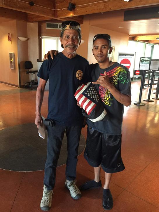 Two men stand close in an airport terminal, displaying an American flag pouch and sharing smiles.