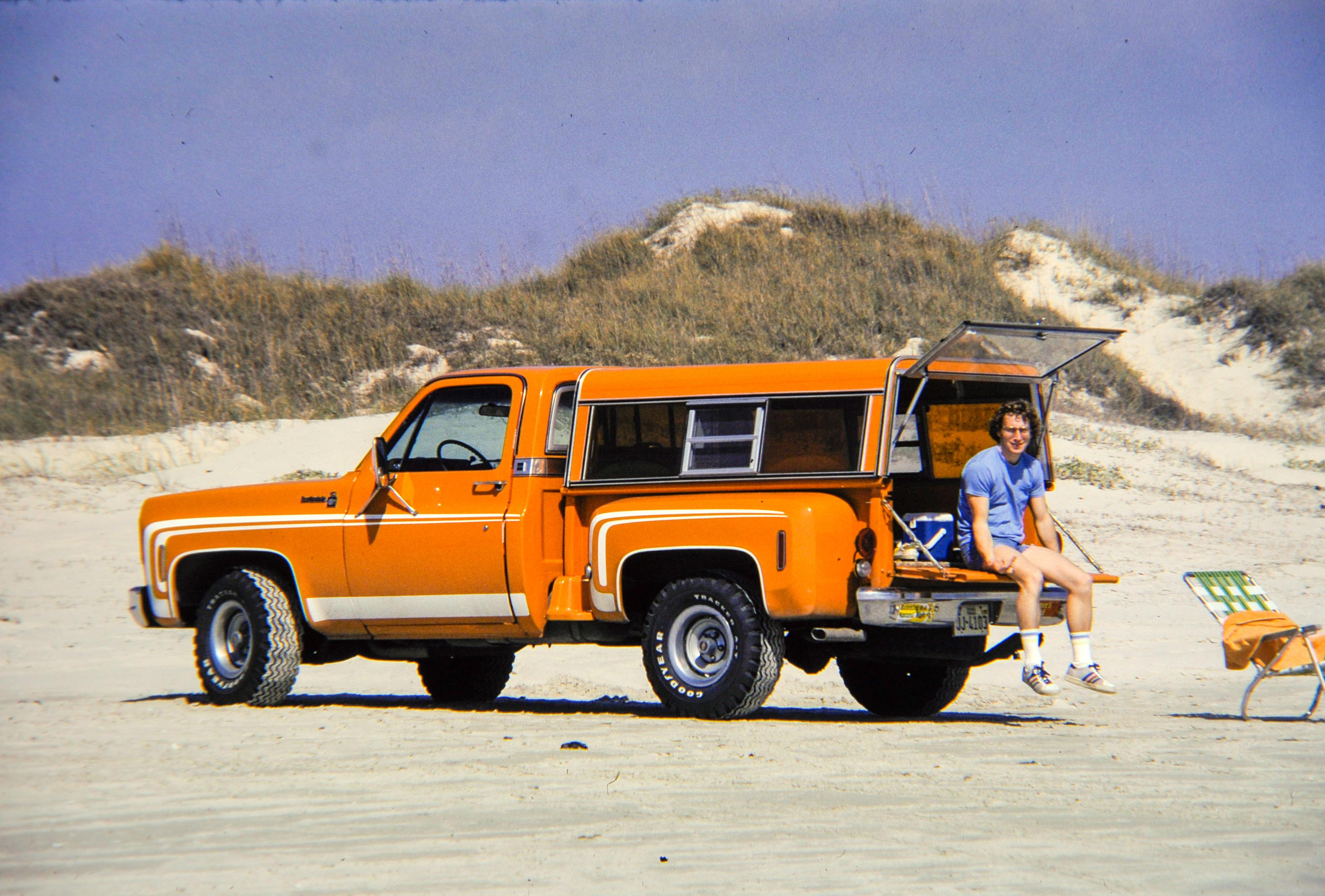 A vintage orange truck parks on the beach while a man relaxes nearby, basking in the sun.