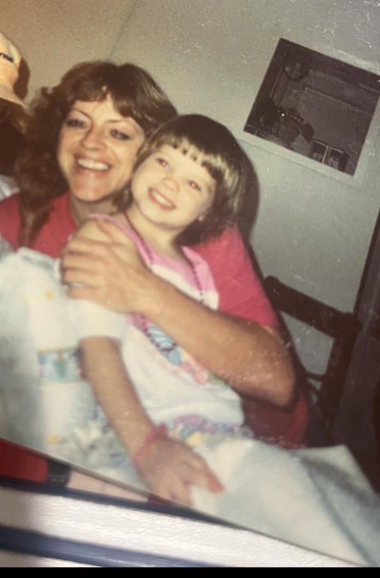 A mother embraces her young daughter, both smiling happily in a cozy home setting.