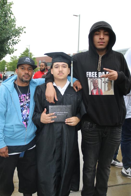 A proud graduate stands with two family members, celebrating a significant achievement outdoors.