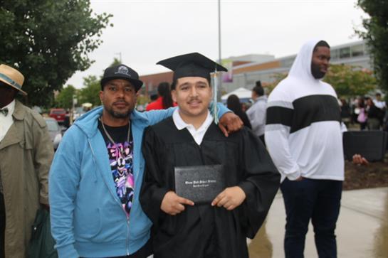 A proud graduate stands with a family member holding a diploma outside a school.