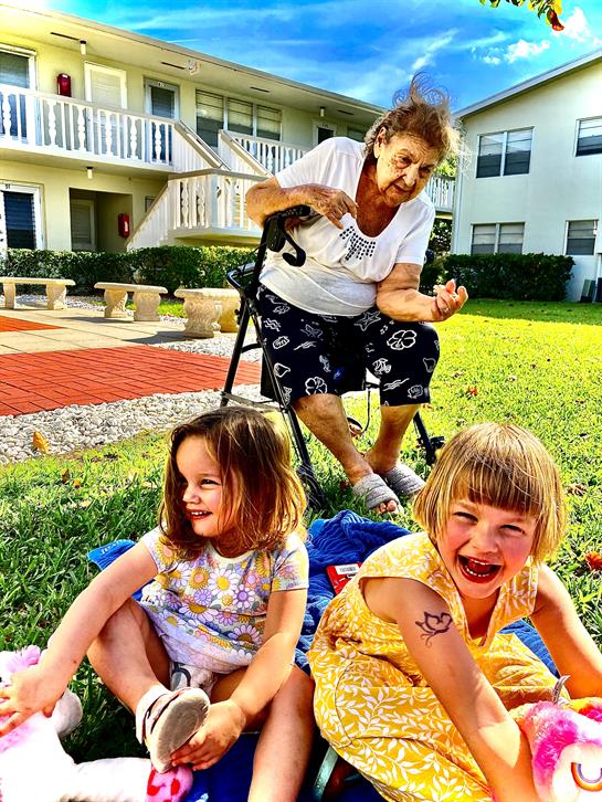 Two girls laugh while playing with stuffed animals near a woman in a wheelchair during the day.