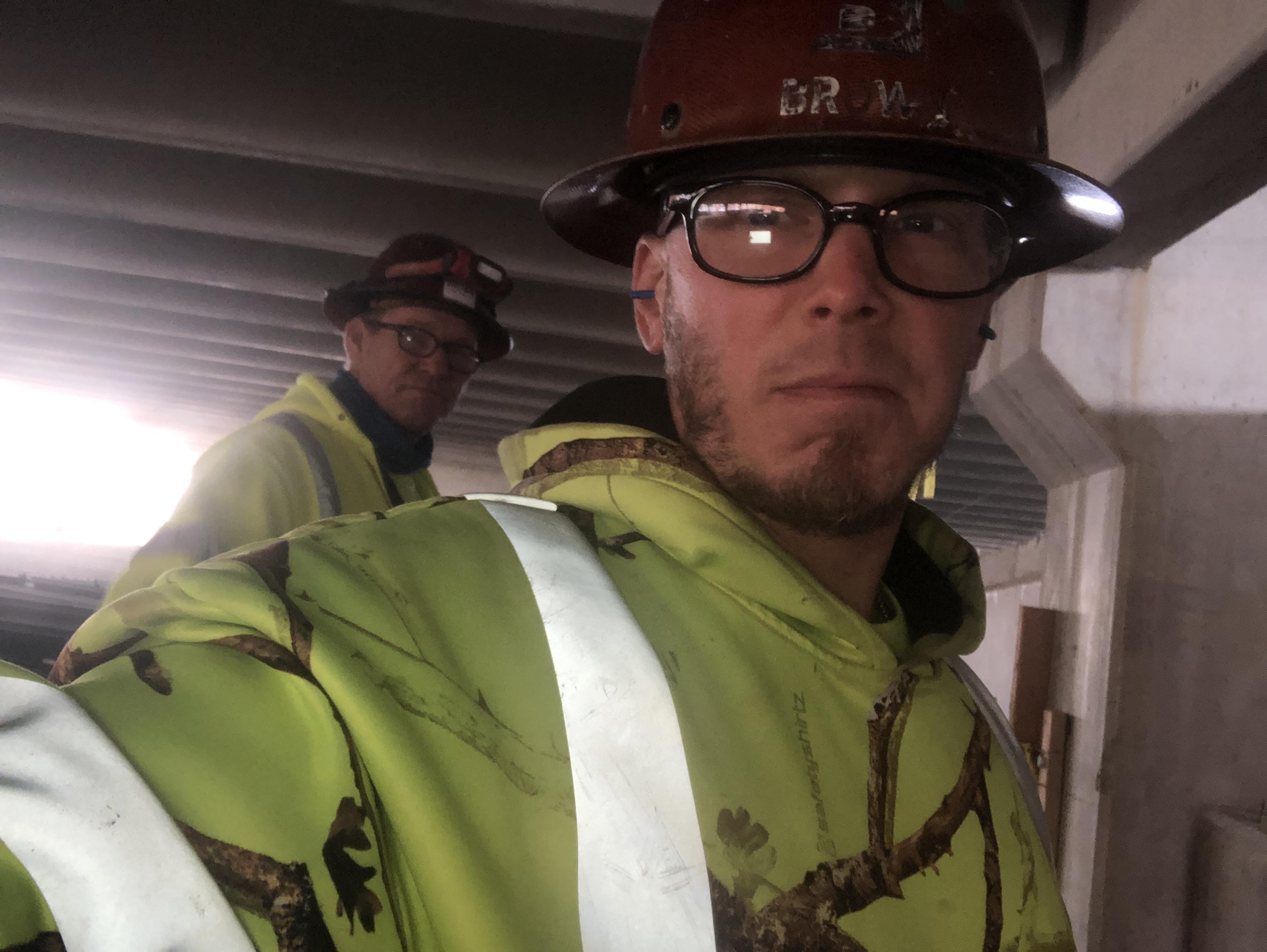 Two construction workers wear safety gear and collaborate inside a partially built structure.