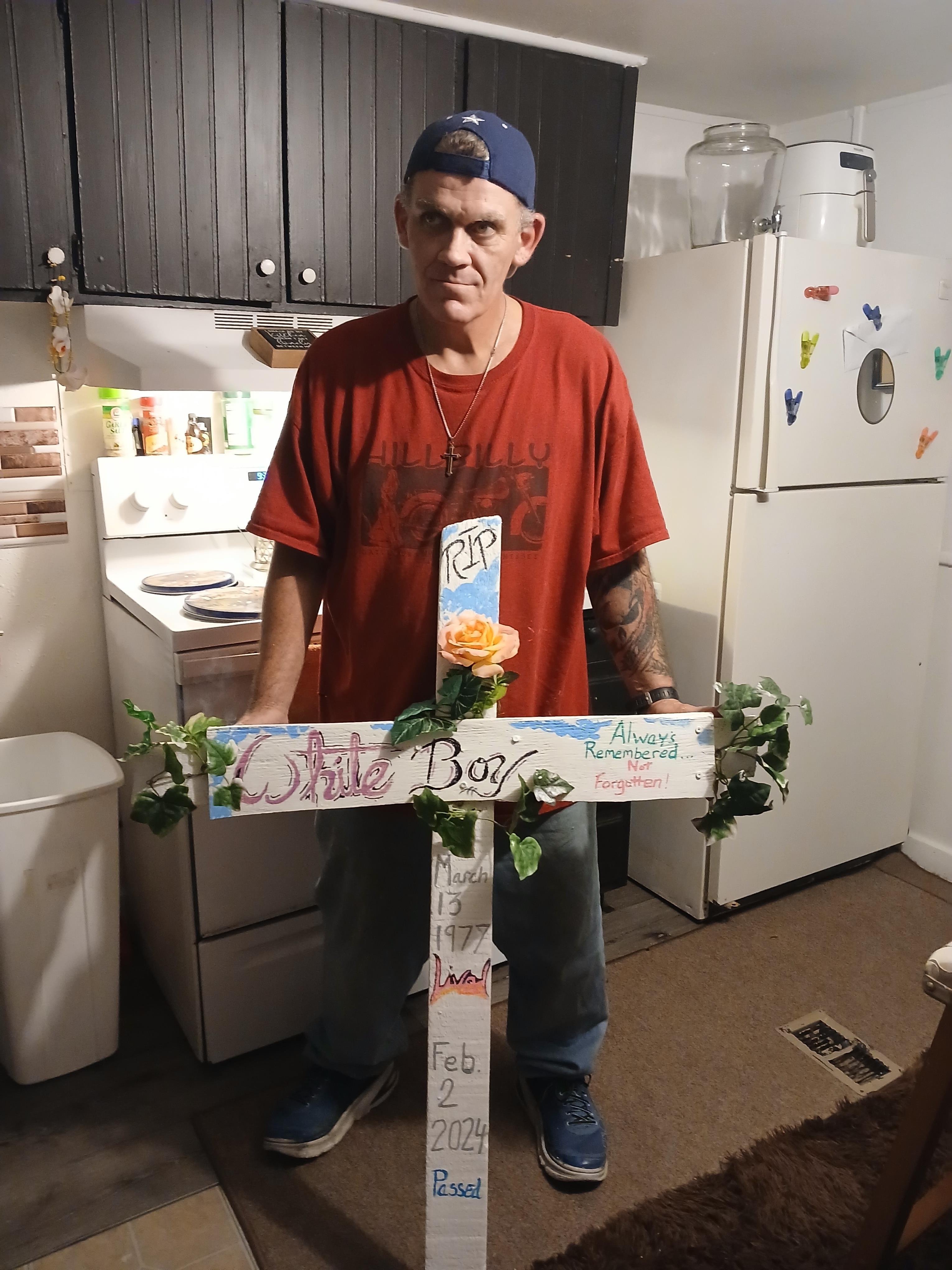 A man stands in a kitchen, displaying a memorial cross adorned with flowers and inscriptions.
