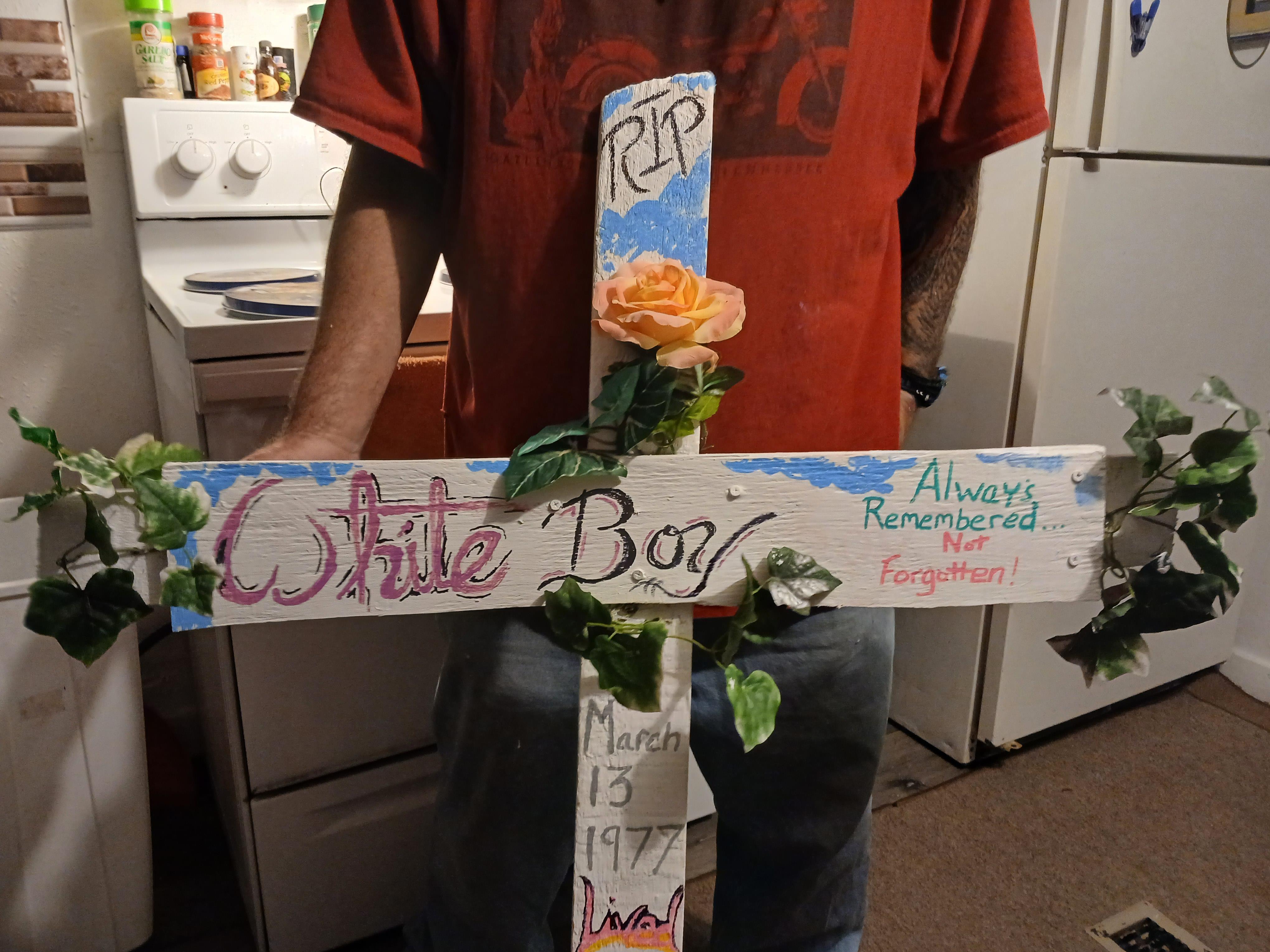 A person stands holding a decorated memorial cross featuring flowers and personal messages.