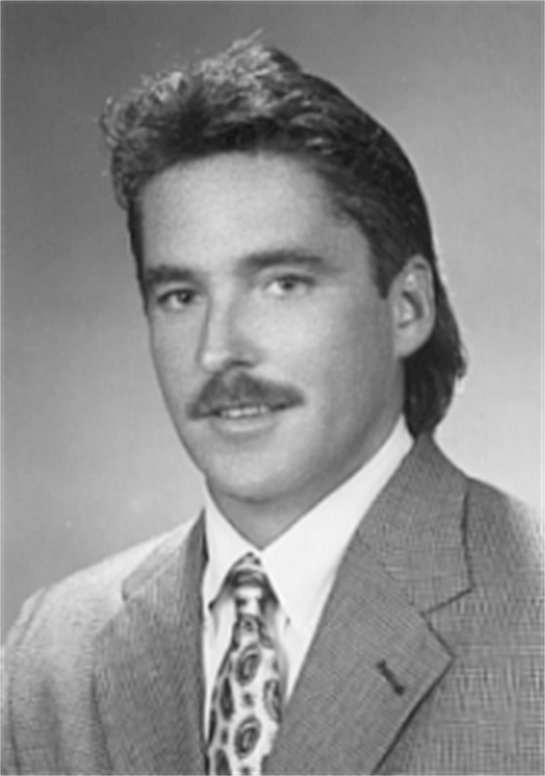 A man with a mustache wearing a gray suit and patterned tie looks confidently at the camera.