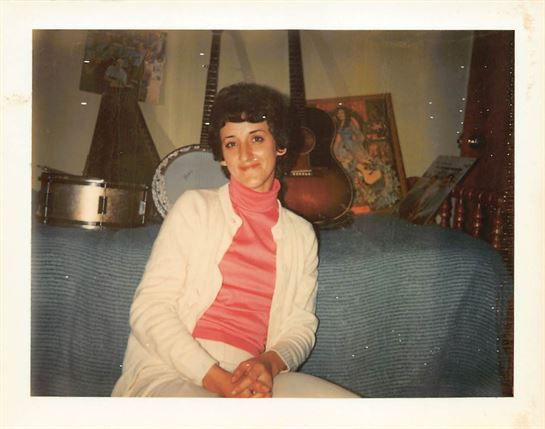 A young woman in a pink sweater poses on a blue couch with musical instruments nearby.
