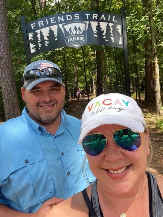 Man and woman smile for a selfie at Friends Trail, enjoying a sunny day in the forest.