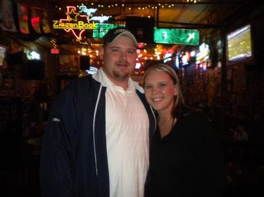 Smiling couple stands together in a vibrant bar with colorful lights and screens.