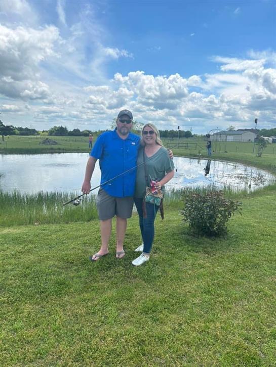 A couple stands together, smiling while holding fishing gear near a calm pond with cloudy skies.