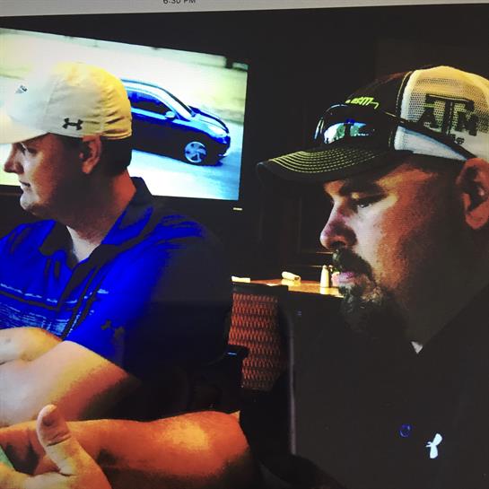 Two men sit at a table, focused on a conversation while a car is shown behind them.