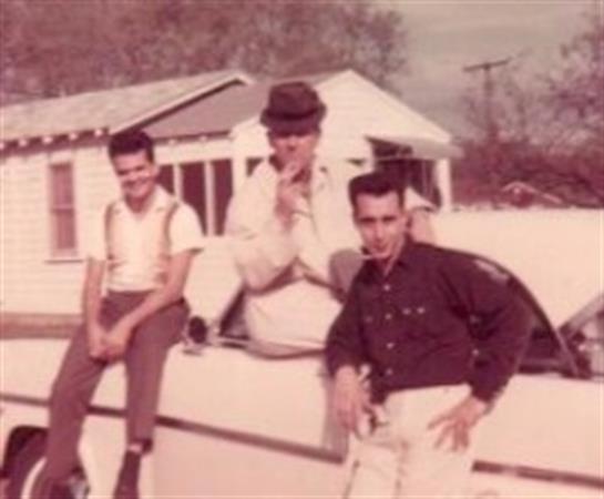 Three men relaxed at a vintage car with an old house in the background during the 1960s.